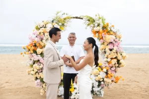 A bride and groom stand on a Bali beach, exchanging rings as part of their wedding vows in Nusa Dua.