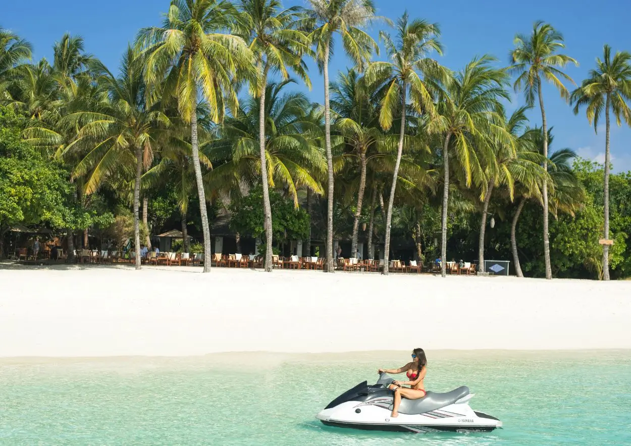 A woman enjoying a thrilling ride on a jet ski along a sandy beach, surrounded by water and a bright sky.